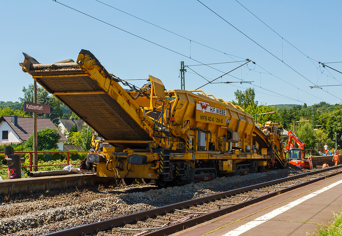 Baustelle im Bahnhof Katzenfurt (Lahn-Dill-Kreis) am 20.07.2013: Der MFS 40/4-B-ZW (mit Bagger) der H. F. Wiebe, Schweres Nebenfahrzeug 90 80 9052 001-1 D-GBM (ex 97 19 13 501 57-5) ist im Einsatz, der integrierte Bagger ist beim Aushub des Oberbaumaterials (Schotter und Planum).

Der MFS 40/4-B-ZW wurden 2009 von Plasser & Theurer unter der Fabriknummer 5479 gebaut und an die Wiebe Gleisbaumaschinen GmbH (Achim) geliefert.

Die MSF-ZW können sich durch seine beiden selbstständig angetriebenen Raupenfahrwerke im gleislosen Baustellenbereich fortbewegen, bzw. durch seine beiden Laufdrehgestelle, vorausgesetzt er besitzt vor sich einen MFS oder einen Schutzwagen mit Auflagerbock für vorragendes Schwenkband, in Züge eingestellt werde

Die beiden MFS 40/4-ZW sind für den gleislosen Einsatz konzipiert. Der MFS 40/4-ZW-B ist, im Bereich des Beladebandes, mit einem Bagger ausgerüstet, mit dessen Hilfe das vorhandene Planum/Aushub aufgenommen und über das Förderband in den Bunker befördert und gespeichert wird. Ist der Bunker fast voll, fährt der ebenfalls mit Raupenfahrwerken ausgestatteten MFS 40/4-ZW-A heran und der Aushub wird an ihn übergeben. Der MFS 40/4-ZW-B kann kontinuierlich weiter arbeiten, während nun der MFS 40/4-ZW-A, in der Zwischenzeit das Material auf im Gleis stehende gleisgebundene MFS, bringt und an diese übergibt. Die MFS sind mit einer eigenen Energieversorgungseinheit ausgestattet, wodurch ein individueller, also voneinander unabhängiger, Einsatz gewährleistet ist.  

TECHNISCHE DATEN des MFS 40/4-ZW-B:
Hersteller: Plasser & Theurer, Fabriknummer 5479 (2009)
Spurweite: 1.435 mm (Normalspur) / oder auf Raupenfahrwerke
Gesamtlänge: 23.920 mm (über alles) 
Länge über Puffer: 19.900 mm
Drehzapfenabstand: 13.000 mm
Breite: 3.150 mm
Höhe: 4.600 mm
Eigengewicht: 72.800 kg
Gesamtgewicht auf Drehgestell: 72.800 kg (leer)
Gesamtgewicht auf Raupe beladen: 128 t
Eigenfahrgeschwindigkeit auf Raupen: bis 3 km/h
Max. Siloinhalt: 25 m³
Förderleistung: 500 m³/h
Kleinster befahrbarer Gleisbogen: R 150 m
Dieselmotor: wassergekühlter 6-Zylinder Reihenmotoren mit Common-Rail-Einspritzsystem, Turboaufladung und Ladeluftkühlung, vom Typ Deutz TCD 2013 L06 2V mit einer Leistung von 160 kW  
Höchstgeschwindigkeit im Zugverband: 100 km/h (geschleppt)
