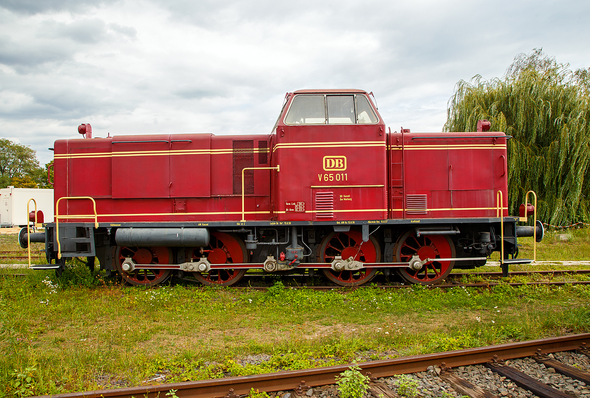 Die DB V 65 011, ex DB 265 011-7, am 04.09.2020 im DB Museum Koblenz-Lützel.

Die V 65 wurde 1955 bei MaK (Maschinenbau Kiel) unter der Fabriknummer 600014 gebaut und als V 65 011 an die Deutsche Bundesbahn (DB) geliefert. Die Umzeichnung in DB 265 011-7 erfolgte 1968, die Ausmusterung bei der DB 1977. Seit 2011 ist sie nun hier im DB Museum Koblenz.

Für den Einsatz auf Nebenbahnen wurde die aus der zum ersten Typenprogamm von MaK gehörigen 600 D die V 65 der DB entwickelt und in 15 Exemplaren an die DB geliefert. Neben der etwas höheren Motorleistung unterscheidet sie sich in diversen Details von der 600 D. Äußerlich am auffälligsten sind die glatten Seiten der Motorhaube, die waagerechten Lamellen des Frontlüfters und die freistehenden Lampen. Ebenfalls neu ist der mit Koks betriebene Dofa-Ofen zum Vorwärmen und Warmhalten.

Nachdem ein Weiterbau der V 65 zugunsten der V 100.10 aufgegeben wurde, kam diese Splittergattung bis 1964 zunächst im Nebenbahndienst im Raum Marburg/Lahn zum Einsatz, wobei neben Güterzügen auch der Personenverkehr auf dem Dienstplan stand. Anschließend gelangten die Loks in den Norden und verdiente ihr Gnadenbrot im Rangierdienst in Hamburg-Altona, Hamburg-Harburg und Puttgarden.

Die Technik der V65
Die Lok ist auch technisch interessant: Erst mit der V16 wurde es kurz vor dem Krieg möglich, die Kraft des Motors hydraulisch zu übertragen. Im Gegensatz zur Dampfmaschine kann der Dieselmotor nicht aus dem Stand ziehen. Eine Kupplung wie im KFZ würde die Kräfte und Gewichte der Eisenbahn nicht überleben. Während des Krieges wurden nur Rangierdieselloks für die Wehrmacht in dieser Technik gebaut. Nach dem Krieg war die V 65 die erste hydraulische Streckendiesellok. Als Antriebsmaschine dient ein langsam laufender MaK- 6-Zylinder- Schiffsdiesel vom Typ MS 301 C mit 72 Liter Hubraum. Das hört man besonders beim Beschleunigen und Starten der Lok. Vom Strömungsgetriebe wird die Kraft auf eine Blindwelle übertragen und gelangt von dort mit Treibstangen auf die vier Achsen- wie bei der Dampflok
Der Stangenantrieb war die Ursache, dass nur 15 Exemplare gebaut wurden. Für höhere Geschwindigkeiten hat man Dampfloks größere Räder gegeben, um die Drehzahl zu begrenzen. Man stelle sich einmal die V65 mit 2m -Rädern der BR 01 Schnellzugdampflok vor.
Ein großer, langsam laufender Dieselmotor darf nicht einfach kalt gestartet werden. Der Verschleiß wäre enorm. So wird die Lok ca. zwei Stunden “vorgeheizt”, ähnlich einer Dampflok. Nur wird hier nicht Kesselwasser erhitzt, sondern das Motoröl. Ab einer Temperatur von etwa 40° C kann der Motor gestartet werden.

Für 72 Liter Hubraum gibt es keinen Elektrostarter. Deshalb wird der Motor mit Druckluft von 30 bar gestartet. Das ist ein akustisches Schauspiel.

Die Kraftübertragung von der Blindwelle zu den Achsen über Kuppelstangen stellt den technischen Übergang von der Dampflok zur modernen Lok dar. Auch viele Ellok Typen aus der Länderbahnepoche zeigten diese archaische Technik. Damit war allerdings die Höchstgeschwindigkeit begrenzt. Man konnte nicht die Räder vergrößern, wie bei Dampflokomotiven. Das war einer der Hauptgründe für die DB, sich bei Streckenlokomotiven für den Antrieb über Drehgestelle zu entscheiden.

Im Gegensatz zu späteren Lieferungen an Privatbahnen, hatten die DB- Lok freistehende Laternen, wie Dampflokomotiven

Bis 1980 wurde die gesamte Baureihe ausgemustert, nachdem vor allem die Gewerkschaften auf eine vorzeitige Ausmusterung drängten: Durch den langsam laufenden MaK-Motor soll sich im Führerhaus regelmäßig eine beträchtliche Hitze entwickelt haben.

Von den 15 gebauten Loks blieben mit V 65 001 und V 65 011 nur zwei in Deutschland erhalten. Die ehemalige V 65 004 hingegen geht in Italien einer ungewissen Zukunft entgegen.

Technische Daten:
Spurweite: 1435 mm
Achsfolge: D
Länge über Puffer: 10.740 mm
Achsstand: 5.800 mm
größte Breite: 3.100 mm
größte Höhe über Schienenoberkante: 4.150 mm
Raddurchmesser neu: 1.250 mm
kleinster befahrbarer Gleisbogen: 50 m
Dienstgewicht: 54 t
Kraftstoffvorrat: 1.130 l
Heizbrennstoff Koks: 100 Kg

Motor : MaK- 6-Zylinder- Viertakt-Schiffsdiesel vom Typ MS 301 C
Leistung: 650 PS (479 kW)
Drehzahl: 750 U/min
Hubraum: 72 Liter

Getriebe: Voith Typ L 37 z
Höchstgeschwindigkeit 80 km/h 