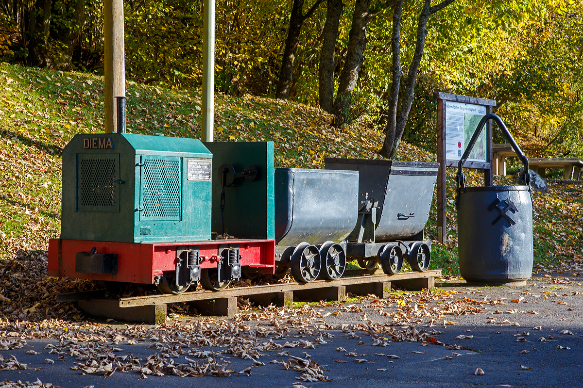 Die DIEMA Feldbahnlok vom Typ DL 6, Fabriknummer 1983, Baujahr 1956, Leistung 18 KW, Gewicht 1.115 kg, von 700 auf 600 mm um gespurt, als Denkmallok am 28.10.2021 mit zwei Grubenwagen beim Wodanstolln in Neunkirchen-Salchendorf. Wobei eigentlich ganz stilecht bzw. geschichtlich richtig ist dieses Denkmal nicht, denn die Lok ist eine Feldbahnlok. Bei den Siegerlnder Erzgruben kamen aber Gubenloks zum Einsatz. 

Die DL 6 der Diepholzer Maschinenfabrik Fritz Schttler (Diema) ist eine Diesellokomotive fr Feldbahnen. Mit einem Gewicht von etwa 1 t, einer Lnge von ca. 1,7 m und einem Achsstand von 0,5 m gehrt sie zu den kleinsten Lokomotiven berhaupt. Die DL 6 entstand 1955 als kleinere und leistungsschwchere Version der DL 8. Insgesamt wurden 220 Exemplare gebaut, womit die DL 6 zu den erfolgreichsten Diema-Konstruktionen gehrt. Eingesetzt wurden die Lokomotiven vor allem von kleinen Torfabbaubetrieben. Die letzte Lokomotive wurde im September 1970 ausgeliefert.

Die Lokomotiven sind auf einem geschweiten Auenrahmen aufgebaut. Die Breite des Rahmens hing von der Spurweite ab, insbesondere gab es eine Version fr die in den Niederlanden verbreitete 700-mm-Spur, zu diese die Lok hier gehrt. In der DL 6 kommen luftgekhlte Einzylinder-Dieselmotoren zum Einsatz. Die Leistungsbertragung erfolgt ber einen Keilriemen vom Motor auf ein mechanisches Zweiganggetriebe und von dort aus ber Rollenketten auf die beiden Achsen. Diese waren zunchst ber Evolutfedern abgesttzt, spter wurden Schraubenfedern eingebaut.

Der Fahrersitz ist, wie bei Lokomotiven dieser Kategorie blich, quer zur Fahrtrichtung eingebaut, so dass der Fahrer seine Position bei Fahrtrichtungswechseln nicht ndern muss. Motor und Getriebe nebst Schalthebeln befinden sich dabei rechts vom Fahrer, der Bremshebel links. Wobei bei dieser Lok der Fahrersitz fehlt.
