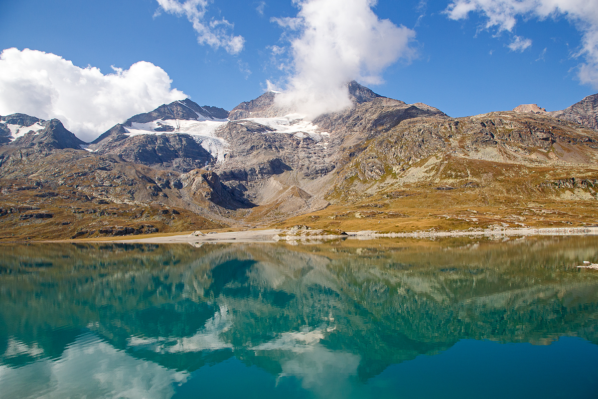 Nun erreichen wir am 06.09.2021den Lago Bianco auf einer Höhe von 2.223 Metern über Meer. 

Hier oben bei der Staumauer befindet sich auch zugleich die Wasserscheide zwischen Donau und Po. In Richtung Süden (Tirano) fließ das Wasser in den Po und somit ins Mittelmeer, in Richtung Norden (St. Moritz) fließ das Wasser über den Inn in die Donau und somit ins Schwarze Meer.

Der Lago Bianco ist ein Stausee am Berninapass zwischen dem Valposchiavo und dem obersten Seitental des Engadins, dem Val Bernina, in der Schweiz. Er liegt damit auf der Grenze zwischen den Bernina- (westlich) und Livigno-Alpen (östlich). Früher bestanden im Gebiet des heutigen Stausees zwei natürlichen Seen, der größere Lago Bianco, , und der kleinere südlich anschließende Lago della Scala. In den Lago Bianco mündeten mehrere kleine Bergbäche von den Höhen der Umgebung. Die beiden Gewichtsstaumauern Scala (Südseite) und Arlas (Nordseite) wurden zwischen 1910–1911 errichtet, so dass sich ein Stausee mit einem Volumen von 18,6 Mio. m³ bildete.

Unmittelbar am Nordende des Lago Bianco liegt die kontinentale Wasserscheide zwischen den Einzugsgebieten des Po und des Inn-Donau-Flusssystems. Wenige Meter nördlich des Lago Bianco liegt der Lago Nero (Schwarzsee) am Oberlauf des Berninabaches, dem folgt dann der kleinere Lago Pitschen (Pitschensee).