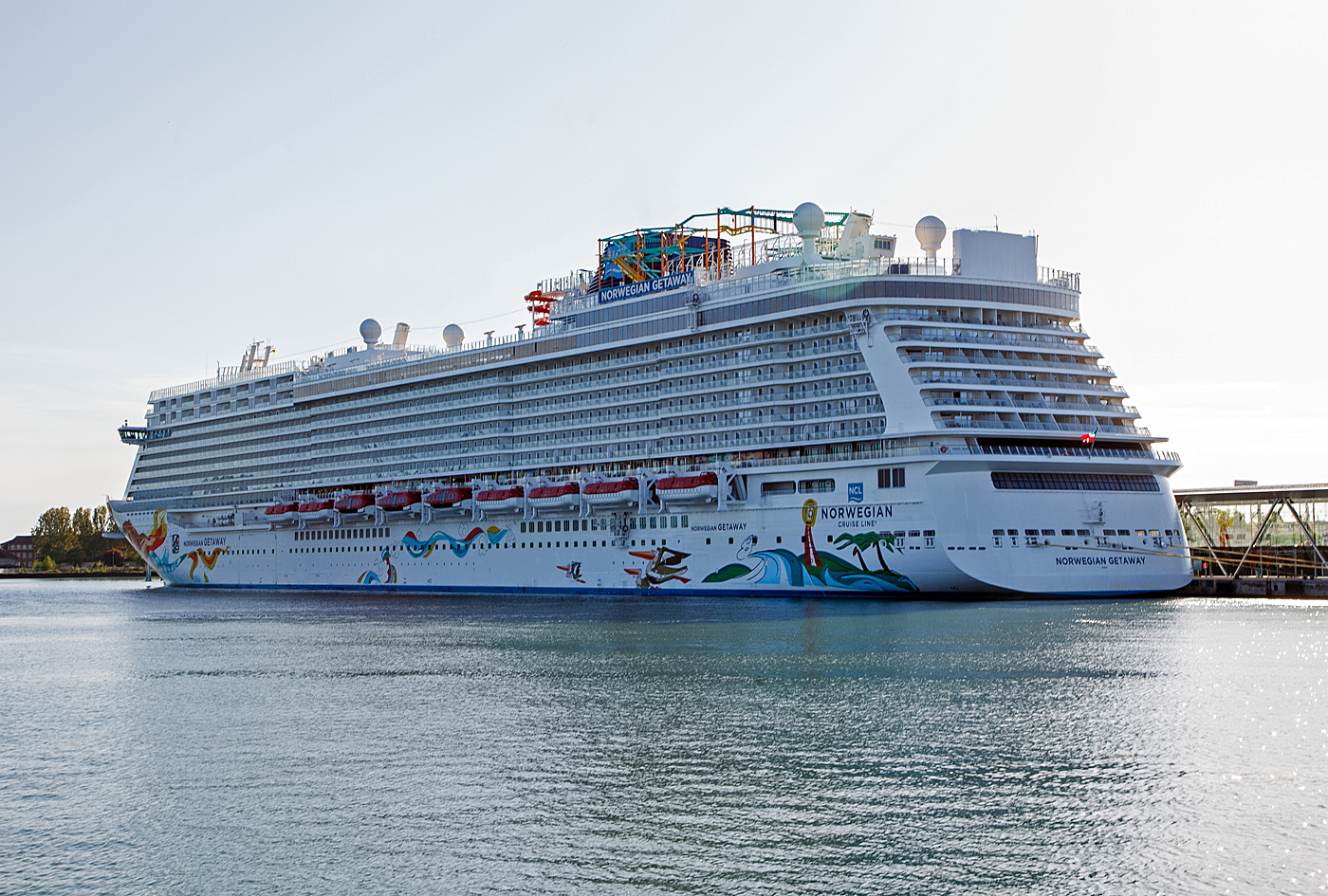 Das gewaltige Kreuzfahrtschiff NORWEGIAN GETAWAY der US-amerikanischen Reederei Norwegian Cruise Line Corporation Ltd. (NCL) liegt am 15 Mai 2022 am Warnemünde Passagierkai, Pier 8. Lieder hier etwas im Gegenlicht.

Wenn man bedenkt dass das Warnemünde Cruise Center für einen Passagierwechsel von bis zu 2.500 Personen am Tag ausgelegt ist, würde es bei diesem Schiff  mit bis zu 4.000 Passagier alleine 1 1/2 Tage brauchen um die kompletten Passagier zu wechseln. Schlimmer wäre aber solch ein Schiff kommt mal in Seenot, wie lange bracht es da um die fast 6.000 Menschen (Passagiere und Besatzungen) sicher von Bord zu bekommen. Da machen wir doch lieber Schienen-Kreuzfahrten. 

TECHNISCHE DATEN der Norwegian Getaway:
Flagge: Bahamas
Bauwerft: Meyer Werft GmbH Papenburger, Deutschland (Baunummer S.692) 
Baureihe: Breakaway-Klasse
Kiellegung / Stapellauf / Übernahme: 30.10,2012 / 02.11. 2013 / 10.01.2014	
Vermessung: 145.655 BRZ
Länge: 324 m
Breite auf Spanten: 39,7 m (über alles 52 m)
Anzahl der Decks: 18
Anzahl der Passagierkabinen: 2.015
Passagiere: 3.963
Besatzung: ca. 1646
Energieversorgung:4 MAN-Dieselgeneratoren
Propeller:	2 × Azipod
Maschinenleistung gesamt: 67.200 kW (84.840 PS)
Antriebsleistung (Dieselelektrischer Antrieb): 42. 000 kW (42 MW)
Max. Geschwindigkeit:  21,5 kn (40 km/h)
Anzahl der Restaurants: 21
Anzahl der Pools: 3

Quellen: Meyer Werft, NCL, Rostock PORT/ Rostock.de, Wikipedia
