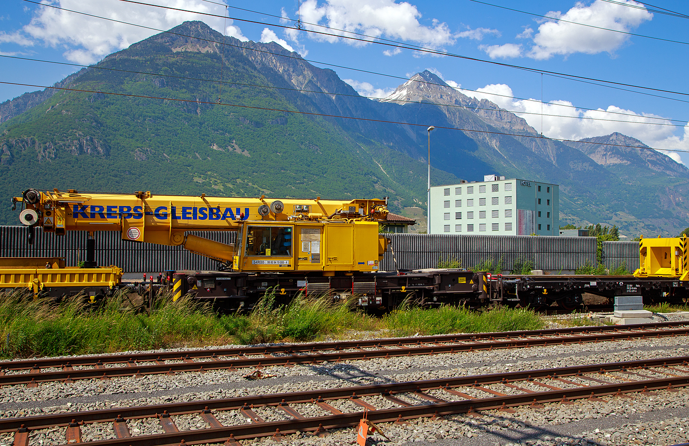 Der 130 t Eisenbahnkran D-KREBS 99 80 9419 008-4, ex 97 82 54 501 17-1, ein GOTTWALD GS 130.06 TT Gleisbauschienenkran der Krebs GmbH & Co. KG, Bingen am Rhein (D) / Bern (CH) ist am 26.05. 2023 beim Bahnhof Martigny, in Transportstellung mit dem Gegenlastwagen 33 88 3936 639-4 B-Krebs der Gattung Rels und dem Schutzwagen auslegerseitig 99 80 9383 003-7 D-Krebs der Gattung Rgs, abgestellt.

Der Kran wurde 2001von GOTTWALD (Mannesmann Dematic) in D�sseldorf unter der Fabriknummer 846 375gebaut.

Der GS 130.06 TT ist eine Weiterentwicklung des GS 80.08 T. Folgende Verbesserungen wurden vorgenommen, die den Einsatzbereich des Kranes nochmals erweitern:
• Der Einsatz eines dreiteiligen Auslegers (ein Grundschuss und zwei Teleskopsch�sse) erm�glicht eine wesentliche Erweiterung des Arbeitsbereiches. Durch die k�rzere Bauform des Auslegers beginnt der verf�gbare Bereich horizontal bereits 3,0 m vor Puffer mit einer Tragf�higkeit von 98,0 t (fr
• eistehend 78,0 t). Damit ist er der einzige am Markt verf�gbare Gleisbaudrehkran, der diese, gerade f�r den Br�ckenbau, wesentliche F�higkeit besitzt.
• Erweiterung des Ausladungsbereiches vor Puffer um 2,0 m auf 15,5 m max. Ausladung.
• Erh�hung der Tragf�higkeit bei max. Ausladung vor Puffer von 15,5 m auf 36 t.
• F�r Schwerlastf�lle ist der Kran mit einem zus�tzlichen Anschlagpunkt versehen, wodurch es jetzt m�glich ist, Lasten von 100 t im 7 m Radius, bis zu einem Schwenkwinkel von 30 � aufzunehmen und zu bewegen.
• Verwendung geschlossener und damit wartungsarmer Fahrgetriebe. Selbstfahrgeschwindigkeit bis max. 25 km/h (zugel. 20 km/h) sind damit m�glich.
• Der Kran verf�gt �ber die M�glichkeit mit nur einer Abst�tzung zu arbeiten, entsprechende Traglasttabellen sind in die Lastmoment�berwachung eingearbeitet. Die Abst�tzungen werden von der Fahrerkabine aus bet�tigt und �berwacht.

Der GS 130.06 TT verf�gt selbstverst�ndlich �ber alle weiteren Ausstattungen, die bereits vom GS 80.08 T bekannt sind und diese Krane gerade f�r den Gleis- und Br�ckenbau so vielseitig und sicher einsetzbar machen, sind wie u.a. den automatischen Gleis�berh�hungsausgleich, jetzt aber bis 180 mm Gleis�berh�hung, die elektronische �berwachung und Steuerung des Lastmomentes unter Einbeziehung der Gegengewichts- und Abst�tzposition u.v.a.m. Die �berh�hungsausgleich-Einrichtung stellt den Kran immer waagerecht. Somit entf�llt die Reduzierung der frei stehenden Tragf�higkeiten.

F�r diesen Krane sind entsprechend Einsatzzweck Traversen unterschiedlicher Tragf�higkeit und Abmessungen verf�gbar.
Dieser Kran ist in die Streckenklasse CE/CM4 eingestuft, das bedeutet, er kann in Regelz�gen bis 120 km/h eingestellt werden. Bei �berstellfahrten muss keine BZA beantragt werden (Schwerlast). Dieser Kran hat durch Ablegen vom Gegengewicht (am Gegengewichtwagen) eine Achslast von 13,5 t.

In Transportstellung wird der Kran mit einem vorangestellten 4-achsiger Schutzwagen auslegerseitig, beladen u.a. mit den zugeh. Traversen, und hier in der Transportstellung wird der Kranausleger hydraulisch auf dem Schutzwagen abgest�tzt, sowie dem vierachsigen Gegenlastwagen (ein Drehgestell-Flachwagen), auf dem Wagen liegen die Gegengewichte (Gegenlast) zum Kran. Durch die Abst�tzung wird die Achslast und folglich die Streckenlast (Meterlast) vom Kran reduziert. Zudem kann eine Lastablegung (Gegengewicht) auf Gegengewichtswagen im Kranbetrieb f�r eine Achslastreduzierung erfolgen.

TECHNISCHE DATEN:
Spurweite: 1.435 mm (Normalspur)
Anzahl der Achsen: 8 (in 4 Drehgestelle bzw. 2 Doppeldrehgestelle)
Unterwagenl�nge �ber Puffer: 13.000 mm
Breite in Transportstellung: 3.100 mm
H�he in Transportstellung: 4.250 mm
Drehzapfenabstand: 8.000 mm
Drehzapfenabstand im Doppeldrehgestelle: 2.300 mm
Achsabstand im Einzel-Drehgestell: 1.100 mm
Laufkreisdurchmesser der R�der: 730 mm (neu) / 680 mm (abgenutzt)
Gesamtgewicht  in Transportstellung:108 t (Gegengewicht abgelegt)
Gr��ter Achsdruck in Transportstellung: 13,5 t
Schwenkwinkel des Oberwagens: 30� 
Max. hintere seitlich Ausladung: 2.000 mm
Maximale Ausladung vor Puffer : 18 m
Kleinster befahrbarer Bogenradius: 90 m
H�chstgeschwindigkeit: 20 km/h (eigen / �BB 25 km/h) / 120 km/h (geschleppt)