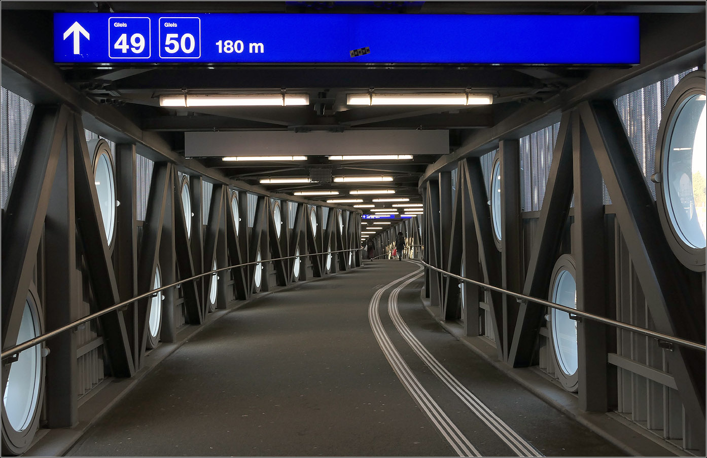 Der Bahnhof Bern - 

Die Fahrgastbrücke zu den 180 Meter entfernten Gleisen 49 und 50 am verlängerten Bahnsteig der Gleise 9 und 10. Der Schwung am Beginn des Stege erklärt sich aus Postion über den Gleisen 8 und 9 im Anschlusse an die 'Welle von Bern.' So musste nicht in die Bahnsteigdächer eingegriffen werden. Im weiteren Verlauf folgt die Brücke dem Bahnsteig, der wiederum eine Krümmung aufweist.

07.03.2025

