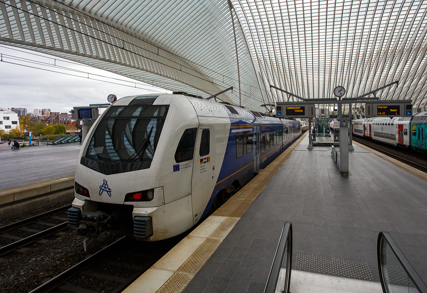 Der dreiteilige, 3-System Stadler FLIRT³ Triebzug 550 (94 84 4 040 550-x NL-AN) der Arriva Personenvervoer Nederland steht am 21 Oktober 2025 im Bahnhof Liège Guillemins (Lüttich-Guillemins), als Dreiländerzug / Drielandentrein / Train des trois pays bzw. RE18 / S 43 (LIMAX - Liège-Maastricht-Aachen-Express (Lüttich über Maastricht nach Aachen) zur Abfahrt bereit. 

Für den Einsatz in der Provinz Limburg beschaffte die Arriva Nederland 38 Triebzüge des Typs Stadler Flirt 3, die ab 2016 in Betrieb gingen. Zum Einsatz kommen dort 15 zwei- und 13 dreiteilige Fahrzeugkompositionen, sowie 8 dieser dreiteiligen Flirt´s in Mehrsystemausstattung (3-System) die von Arriva NL auf dem grenzüberschreitenden Dreiländerzug zwischen Lüttich über Maastricht nach Aachen eingesetzt werden. Die 8 Mehrsystem-FLIRT wurden 2017 bis 2020 in Dienst gesetzt. 

Im Februar 2016 bestellte Arriva Personenvervoer Nederland BV acht dreiteilige FLIRT-Elektrotriebzüge mit Niederflurtechnik bei Stadler. Die Dreisystemzüge können mit 1,5 kV Gleichstrom, 3 kV Gleichstrom und 15 kV Wechselstrom betrieben werden und verkehren im grenzüberschreitenden Regionalverkehr nach Belgien und Deutschland. Die neueste Generation der dreiteiligen FLIRT-Triebzüge ist für eine Höchstgeschwindigkeit von 160 km/h ausgelegt. Sie bieten 172 Sitzplätze, davon 12 in der ersten Klasse und 13 Klappsitze. Die Triebzüge verfügen über Luftfederung und breite Einstiegstüren. Dank des Niederfluranteils von 90 Prozent ist der Zugang zu den klimatisierten Zügen einfach, und die optimale Raumausnutzung gewährleistet ausreichend Platz für Kinderwagen und Rollstühle.

Der Dreiländerzug, niederländisch Drielandentrein, französisch train des trois pays oder LIMAX (Akronym aus Liège-Maastricht-Aachen-Express) ist eine Bahnverbindung, die von Arriva Personenvervoer Nederland betrieben wird.Sie verläuft von Aachen Hbf über Herzogenrath (Deutschland), Heerlen, Maastricht (Niederlande) und Visé nach Liège-Guillemins (Belgien).

Seit dem 29. Januar 2019 verkehren die Züge halbstündlich zwischen Maastricht und Heerlen sowie stündlich grenzüberschreitend bis Aachen. Inzwischen wurde der Halbstundentakt durch den zweigleisigen Ausbau zwischen Heerlen und Landgraaf bis Herzogenrath ausgeweitet. Seit Dezember 2023 wird ebenfalls Aachen zwei Mal pro Stunde erreicht, was vorher durch Trassenkonflikte mit dem RE 4 nicht möglich war. Die stündliche Verlängerung der Linie nach Lüttich fand am 30. Juni 2024 statt. Ziel ist, die Verbindung Lüttich–Maastricht–Aachen im 30-Minuten-Takt zu bedienen.

Der FLIRT (Akronym für Flinker Leichter Innovativer Regionaltriebzug) basiert auf dem Stadler GTW. Der Hauptunterschied besteht darin, dass die Antriebseinheiten des FLIRT hinter den Führerständen angeordnet sind (anstatt in einem separaten Triebwagen). Dieser Zugtyp zeichnet sich durch helle und geräumige Innenräume aus, die durch den Einsatz von Jacobs-Drehgestellen in Kombination mit breiten, offenen Übergängen und einem Niederfluraufbau ermöglicht werden. Der Niederfluraufbau, zusammen mit einer ausziehbaren Trittstufe, verbessert die Zugzugbenutzung für Menschen mit körperlichen Einschränkungen. Das geringe Gewicht des Zuges in Verbindung mit hohem Drehmoment macht ihn ideal für Züge mit häufigen und kurzen Haltezeiten.

Konstruktion:
Die FLIRT-Züge bestehen hier drei Wagen, die an den Wagenübergängen von Jacobs-Drehgestellen getragen werden. Der Einsatz von Jacobs-Drehgestellen ermöglicht kurze und breite Wagenübergänge und sorgt so für hohe Transparenz. Daher verfügen diese Züge über breite Wagenübergänge und einen Niederflureinstieg, wie er bei U-Bahnen und Straßenbahnen üblich ist. Die gute Sicht im gesamten Zug, kombiniert mit den Kameras, soll ein Gefühl der Sicherheit vermitteln. Die Bodenhöhe von 780 mm entspricht der der meisten Bahnsteige. Bis zu drei Züge können im Verbund verkehren.

Im Innenraum gibt es keine Trennwände und Türen; lediglich die erste und zweite Klasse sind durch Glastüren und Trennwände voneinander getrennt. Die Züge sind unter anderem mit Klimaanlage, einem Fahrgastinformationssystem mit TFT-Displays und automatischer Ansage, (Sicherheits-)Kameras und einem Notrufsystem ausgestattet, das nach Betätigung der Notbremse einen akustischen Kontakt zum Lokführer ermöglicht.

Interieur/Innenraum:
Fahrgäste haben Zugang zu USB-Ladestationen für mobile Geräte, Steckdosen und kostenlosem WLAN. Die Züge sind zudem mit einem Niederflurzugang, ausziehbaren Trittstufen und einer rollstuhlgerechten Toilette ausgestattet, um das Reisen für Menschen mit körperlichen Einschränkungen zu erleichtern. Diese Toilette verfügt über eine klappbare Wickelauflage. Die Züge bieten Abteile der ersten und zweiten Klasse, einen Salon und einen Fahrradabstellraum. Die Sitze der ersten Klasse (Kiel Match G2) sind grau mit orangefarbenen Kopfstützen. Die Sitze der zweiten Klasse sind blau-kariert mit dunkelblauen Kopfstützen.

Die 3-System-Züge:
Die Züge sind am neuen Arriva-Logo, den Aufklebern unterhalb der Führerstandsfenster mit dem Text „Kofinanziert von der Europäischen Union“ und den Dreiländerzug-Aufklebern erkennbar. Auf Wunsch Belgiens fehlt an den Führerstands Seiten der, sonst bei Arriva übliche, Limburg-Löwen. Die Züge sind primär für den Schnellzugverkehr konzipiert, sie verfügen über drei Türen pro Seite, viele Sitzplätze, wenige Klappsitze und wenige Fahrrad- oder Rollstuhlplätze. Im internationalen Verkehr werden Reiseinformationen auf Niederländisch, Deutsch und Französisch durchgesagt. Im Mittelteil gibt es zwei querangeordnete Sitzbänke. 

Die 3-System Stadler FLIRT³ (Baureihe IIImcs) erzeugen ein anderes Geräusch als die reine 1,5 kV- Gleichstrom-Triebzüge (Baureihe II und III). Dieses Geräusch wird durch die Traktionsumrichter verursacht, die es dem Triebzug ermöglichen, mit verschiedenen Spannungen zu arbeiten. Diese Umrichter sind von anderer Bauart und nutzen ebenfalls eine Drei-Phasen-Technologie, was ein leichtes Brummen des Triebzugs verursacht. Bei einer Oberleitungsspannung von 1,5 kV unterscheidet sich dieses Geräusch von dem bei 3 kV und 15 kV. Auch bei 1,5 kV ist ein Brummen hörbar, das von einer Drosselspule auf dem Dach stammt.

Technik der 3-System-Züge: 
Die 3-System-Triebzüge verfügen über zwei Stromabnehmer auf dem Mittelwagen, einen für Gleichstrom (1,5 und 3 kV) und einen für Wechselstrom (15 kV). Hinter jedem Führerstand befindet sich ein Stromumrichter (Fabrikat ABB, Type BORDLINE CC1500 MS_15-3-1.5kV_M_1300_070A01), der die Oberleitungsspannung in Drehstrom für die Fahrmotoren umwandelt. Die Umrichter arbeiten mit schnell schaltenden IGBTs; die Schaltfrequenz ist während der Fahrt hörbar. Beide Fahrmotoren (Fabrikat TSA, Type TMF 59-33-4 (600 kW)) eines Drehgestells werden gemeinsam gesteuert. Die Zugkraft der Fahrmotoren wird über ein Zahnradgetriebe mit einem Übersetzungsverhältnis von 1:5,347 auf die Achsen übertragen.

Auslieferung:
Der erste 3-System-Zug (550) wurde am 19. September 2017 über Venlo ausgeliefert. Da ein belgisches Gesetz (von 2014) die Ausstattung neuer Züge im Schienennetz mit dem ETCS-Sicherheitssystem vorschreibt, mussten die IIImcs entsprechend ausgerüstet werden. Dies erfolgte im Zeitraum 2021–2023 bei Stadler Refit in Blerick. Die ersten Testfahrten in Belgien fanden am 1. Juni 2022 statt, die Züge 550 und 555 waren bereits mit ETCS ausgestattet, andere noch nicht. Die endgültige Umrüstung auf ETCS erfolgte 2023, woraufhin Anfang 2024 Testfahrten und die Zulassung für Belgien folgten.

TECHNISCHE DATEN der 3-System Stadler FLIRT³:
Gebaute Anzahl: 8 (dreiteilig)
Nummerierung: 550 bis 557
Hersteller: Stadler Rail
Baujahre: ab 2017
Spurweite: 1.435 mm (Normalspur)
Achsfolge: Bo'2'2'Bo' (8 Achsen, davon 4 angetrieben) 
Länge über Kupplung: 63.200 mm
Fahrzeugbreite: 2.820 mm
Fahrzeughöhe: 4.120 mm
Drehzapfenlabstände: 17.600 mm / 17.500 mm / 17.500 mm / 17.600 mm
Achsabstand im Triebdrehgestell: 2.500 mm
Achsabstand im Laufdrehgestell: 2.700 mm
Treibraddurchmesser: 920 mm (neu)
Laufraddurchmesser: 760 mm (neu) / 690 mm (abgenutzt)
Leergewicht: 121 t
Höchstgeschwindigkeit: 160 km/h (in NL und B planmäßig 140 km/h) / 160 km/h (in D)
Sitzplätze: 12 in 1. Klasse, 147 der 2. Klasse davon 13 Klappsitze
Stehplätze: 161 (4 Personen/m²)
Bodenhöhe: Niederflur 780 mm (90%), Hochflur 1.180 mm
Türbreite: 1.300 mm
Türen je Längsseite: 3
Leistung am Rad: 3.000 kW
Anfahrzugkraft: 200 kN
Anzahl der Traktionsmotoren: 4 TSA Typ TMF 59-33-4 (600 kW)
Übersetzung: 1 : 5,347
Stromsysteme: 1,5 kV DC, 3,0 kV DC (Gleichstrom) und 15 kV 16,7 Hz AC (Wechselstrom)
Beschleunigung bis 54 km/h: 0,95 m/s²
Verzögerung: 1,15 m/s²
Kupplung: Scharfenberg
