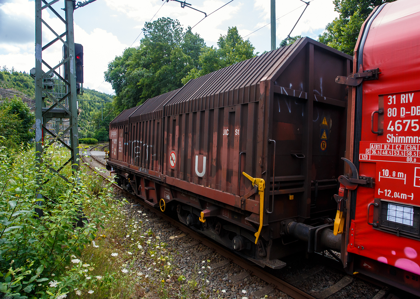 Vierachsiger Drehgestell-Flachwagen mit verschiebbaren Teleskophauben für Coiltransporte, 31 80 4671 011-3 D-DB, der Gattung Shimmns-u 708, der DB Cargo AG, am 16 Juli 2025 im Zugverband (Zug 9921) bei der Zugdurchfahrt in Kirchen (Sieg) in Richtung Siegen bzw. Ziel Kreuztal. 

Der Wagen eignet sich speziell für den Transport witterungsempfindlicher Blechrollen. Er besitzt feste Stirnwände, 5 fest im Untergestell eingebaute Lademulden und drei teleskopartig angeordnete Hauben, die sich zu beiden Wagenenden hin soweit durch Ineinanderschieben öffnen lassen, dass jeweils 2/3 des Wagens freigelegt werden kann. Hierdurch ist eine einfache Beladung des Wagens sowohl von oben als auch von der Seite aus möglich. Der Wagen darf nur mit geschlossenen und verriegelten Hauben verkehren.

Das Untergestell und die festen Stirnwände, die als Abkantrahmenkonstruktion mit Blechbekleidung und hutförmigen Verstärkungsprofilen ausgeführt sind, sowie die fest auf den Außenlangträgern angeordneten drei Laufschienen je Wagenseite bilden den als Schweißkonstruktion ausgeführten Wagenkasten. Das Untergestell mit seinen beiden kräftigen Außenlangträgern besteht aus Walz- und Abkantprofilen. Die Untergestellenden bestehen aus den Hauptquerträgern, den AK-Trägern und den Kopfstücken. Die sattelförmig angeordneten, jeweils mit den Außenlangträgern verschweißten Querträger bilden zusammen mit 6 mm dicken Muldenblechen die Lademulden.

Die teleskopartig ineinander verschiebbaren Stahlhauben bestehen aus einer Hohlprofil-Rahmenkonstruktion und einer mit Sicken versehenen Beblechung. Die Abdichtung der Hauben untereinander und zu den Stirnwänden hin erfolgt über labyrinthartig ausgeführten Umrandungsprofilen. Die Endhauben verriegeln in geschlossener Stellung automatisch. Das Entriegeln der Hauben ist sowohl vom Erdboden wie auch von der Rampe möglich. Die beidseitig an jeder Stirnwand angeordneten Verriegelungseinrichtungen sind durch Wellen miteinander verbunden. Hierdurch ist das Entriegeln von nur einer Wagenseite aus möglich. Jede Haube läuft auf 4 jeweils und mit Abhebesicherungen versehenen Laufwagen mit nadelgelagerten Doppelrollen.

Die Wagen der Bauart Shimmns 708 sind mit 12 verstellbaren Festlegearmen ausgerüstet, die in fest auf den Muldenkappen angeordneten kammartig ausgeführten Halterungen gelagert sind. Die Festlegearme und ihre Anordnung ergeben einen wirksamen Schutz gegen unzulässiges seitliches Verschieben der Coils.

Um unzulässige Radlastabweichungen auszuschließen, ist eine möglichst mittige Verladung der Coils erforderlich. Ein durch alle Mulden in Wagenmitte verlaufender weißer Farbstreifen dient hierzu als Orientierungshilfe.

TECHNISCHE DATEN:
Hersteller: LHB - Linke-Hofmann-Busch GmbH in Salzgitter-Watenstedt
Baujahr: 1972, Fabriknummer 407
Spurweite: 1.435 mm (Normalspur)
Achsanzahl: 4 (in 2 Drehgestelle)
Länge über Puffer: 12.040 mm
Länge der Ladefläche: 10.800 mm
Ladebreite (Mulden): 2.400 mm
Drehzapfenabstand: 7.000 mm
Achsabstand im Drehgestell: 1.800 mm
Laufraddurchmesser: 920 mm (neu)
Eigengewicht: 22.800 kg
Höchstgeschwindigkeit: 100 km/h (beladen) / 120 km/h (leer)
Maximales Ladegewicht: 67,0 t (ab Streckenklasse D4) / 61,0 t bei DB ab CM3
Hier betrug das Ladegewicht laut Laufzettel 65.055 kg
Kleinster bef. Gleisbogenradius: 35 m
Bauart der Bremse: KE-GP (LL)
Bremssohle: IB 116
Handbremse: Ja
Intern. Verwendungsfähigkeit: RIV